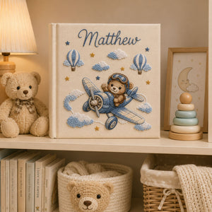 Personalized hand embroidered photo album featuring a teddy bear pilot flying a vintage airplane with clouds hot air balloons and stars on a linen cover