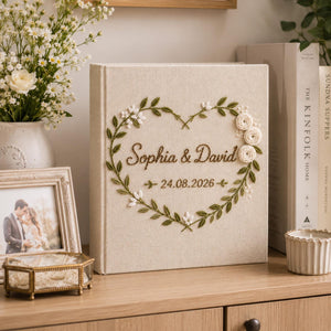 Personalized embroidered wedding photo album with heart-shaped floral wreath, white rose embroidery, botanical greenery, custom couple names, wedding date, and elegant linen hardcover design