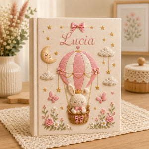 Personalized hand embroidered photo album featuring a crowned bunny in an elegant balloon basket beneath a sleepy crescent moon with hanging stars, butterflies, clouds, flowers, and custom name embroidery on a premium linen cover