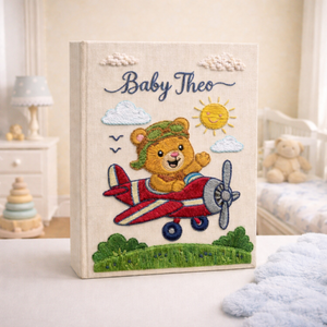 Personalized hand embroidered photo album featuring a teddy bear pilot flying a vintage red airplane beneath clouds and sunshine on a premium linen cover