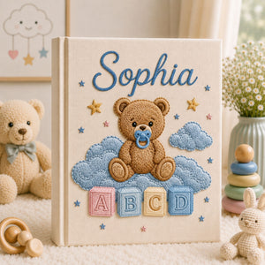 Personalized hand embroidered photo album featuring a teddy bear with a pacifier sitting on clouds above colorful ABC blocks on a linen cover
