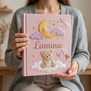 Personalized hand embroidered photo album featuring a teddy bear holding a baby bottle on fluffy clouds beneath a crescent moon with heart balloons and stars on a pink linen cover customized with a baby name