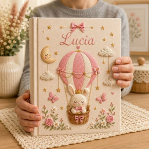 Personalized hand embroidered photo album featuring a crowned bunny in an elegant balloon basket beneath a sleepy crescent moon with hanging stars, butterflies, clouds, flowers, and custom name embroidery on a premium linen cover