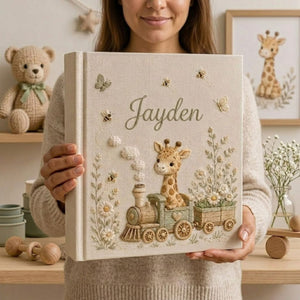Personalized hand embroidered photo album featuring a baby giraffe riding a vintage train surrounded by daisies, butterflies, and honeybees on a premium linen cover customized with a baby name