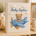 Personalized hand embroidered photo album featuring a teddy bear piloting a blue vintage airplane among fluffy clouds on a premium linen cover customized with a baby name