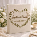 Personalized embroidered wedding photo album with heart-shaped floral wreath, white rose embroidery, botanical greenery, custom couple names, wedding date, and elegant linen hardcover design