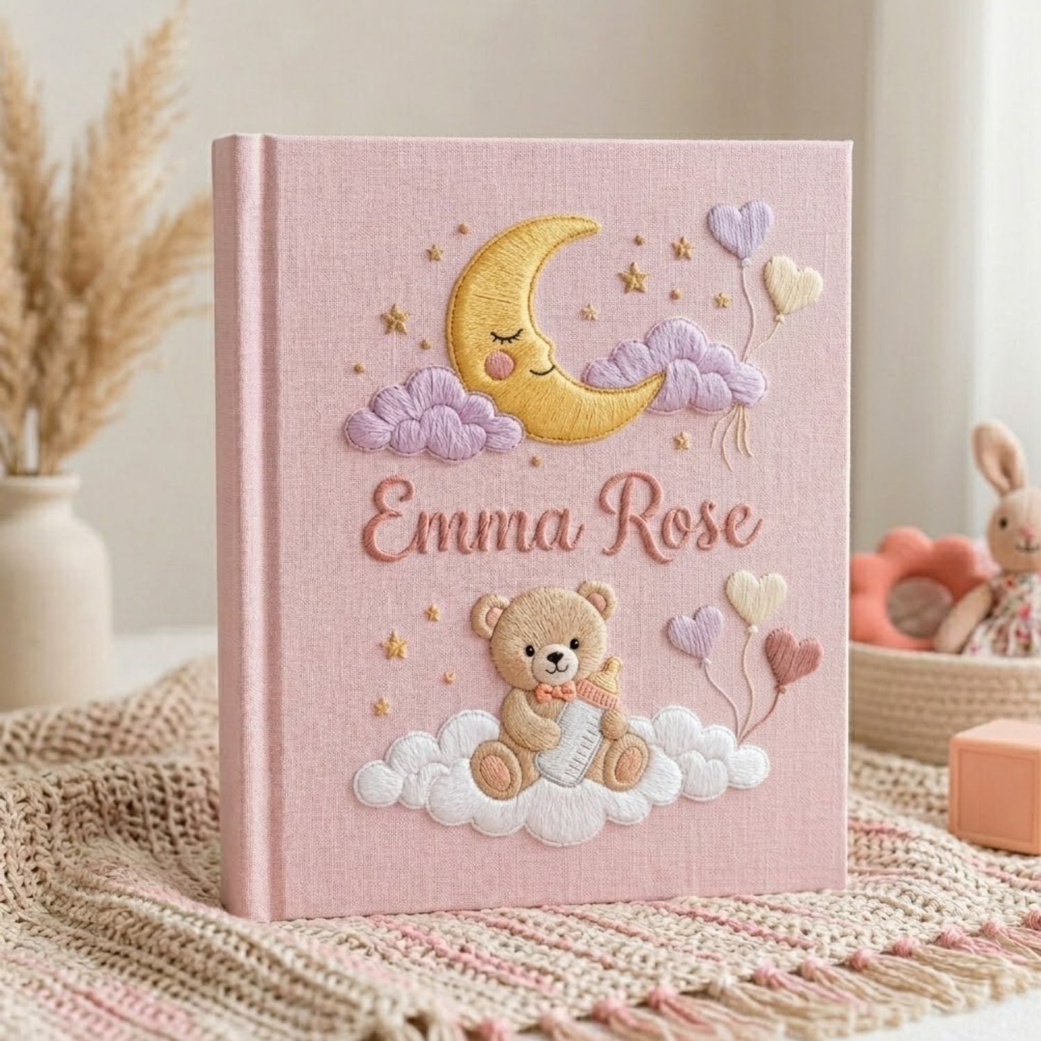 Personalized hand embroidered photo album featuring a teddy bear holding a baby bottle on fluffy clouds beneath a crescent moon with heart balloons and stars on a pink linen cover customized with a baby name