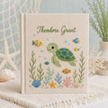 Personalized hand embroidered photo album featuring a cute sea turtle swimming with tropical fish, coral, seashells, and ocean plants on a premium linen cover customized with a baby name