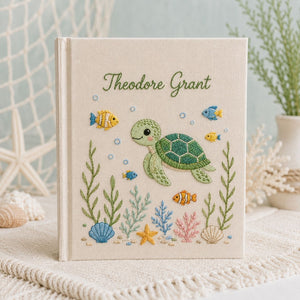 Personalized hand embroidered photo album featuring a cute sea turtle swimming with tropical fish, coral, seashells, and ocean plants on a premium linen cover customized with a baby name