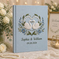 Personalized embroidered wedding photo album with white swans, blue floral heart wreath embroidery, custom couple names, wedding date, and elegant blue linen hardcover design