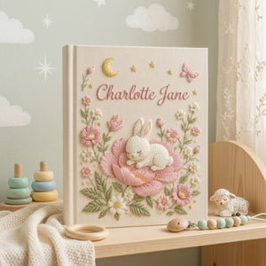 Personalized hand embroidered photo album featuring a sleeping bunny resting on a large pink flower surrounded by blossoms, butterflies, stars, and a crescent moon on a premium linen cover customized with a baby name