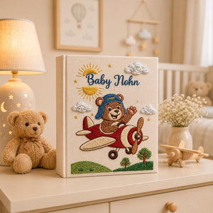 Personalized hand embroidered baby photo album with teddy bear pilot flying a vintage airplane surrounded by clouds and sunshine