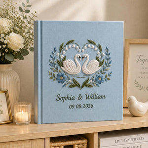 Personalized embroidered wedding photo album with white swans, blue floral heart wreath embroidery, custom couple names, wedding date, and elegant blue linen hardcover design