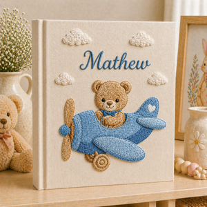 Personalized hand embroidered photo album featuring a teddy bear piloting a blue vintage airplane among fluffy clouds on a premium linen cover customized with a baby name