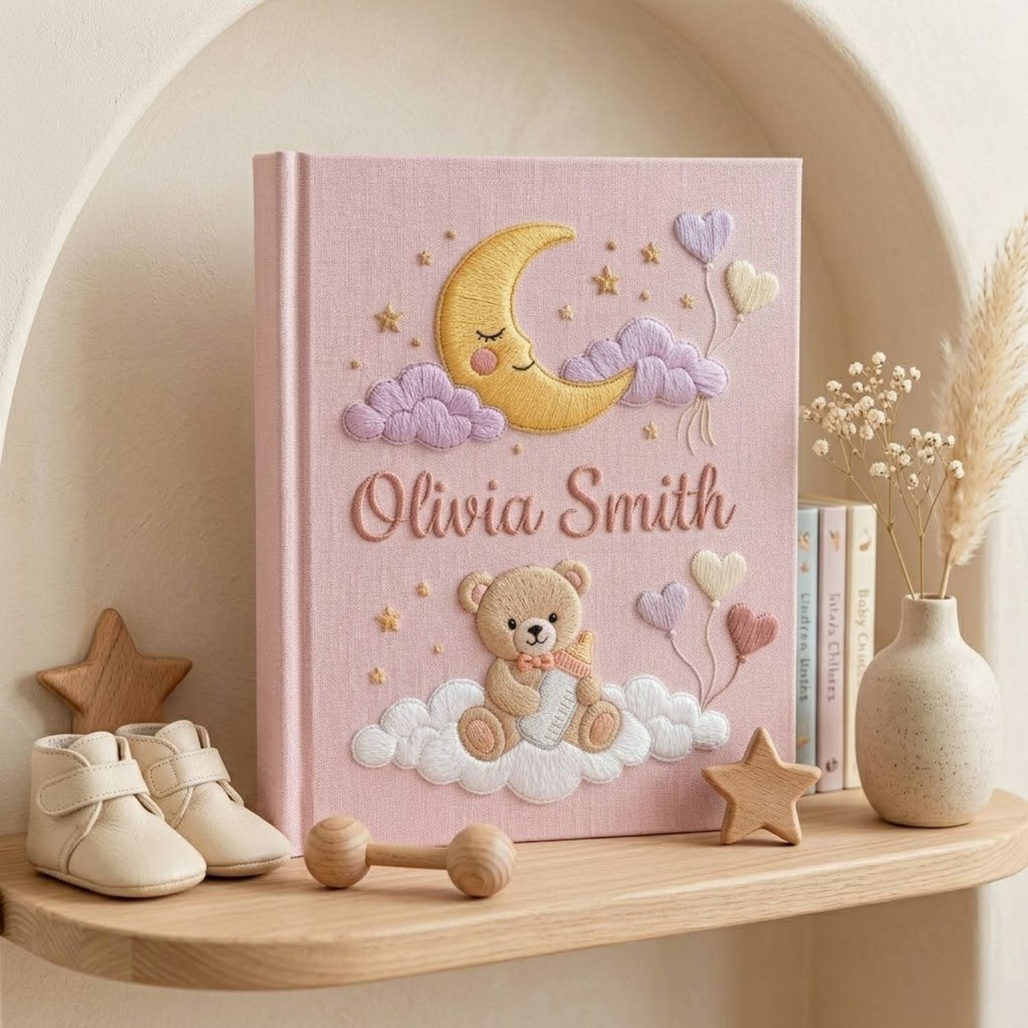 Personalized hand embroidered photo album featuring a teddy bear holding a baby bottle on fluffy clouds beneath a crescent moon with heart balloons and stars on a pink linen cover customized with a baby name