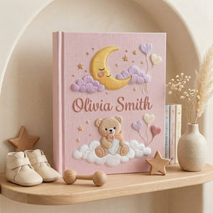 Personalized hand embroidered photo album featuring a teddy bear holding a baby bottle on fluffy clouds beneath a crescent moon with heart balloons and stars on a pink linen cover customized with a baby name