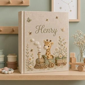 Personalized hand embroidered photo album featuring a baby giraffe riding a vintage train surrounded by daisies, butterflies, and honeybees on a premium linen cover customized with a baby name