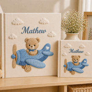 Personalized hand embroidered photo album featuring a teddy bear piloting a blue vintage airplane among fluffy clouds on a premium linen cover customized with a baby name