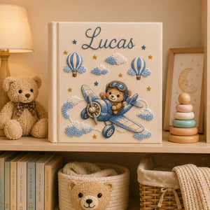 Personalized hand embroidered photo album featuring a teddy bear pilot flying a vintage airplane with clouds hot air balloons and stars on a linen cover