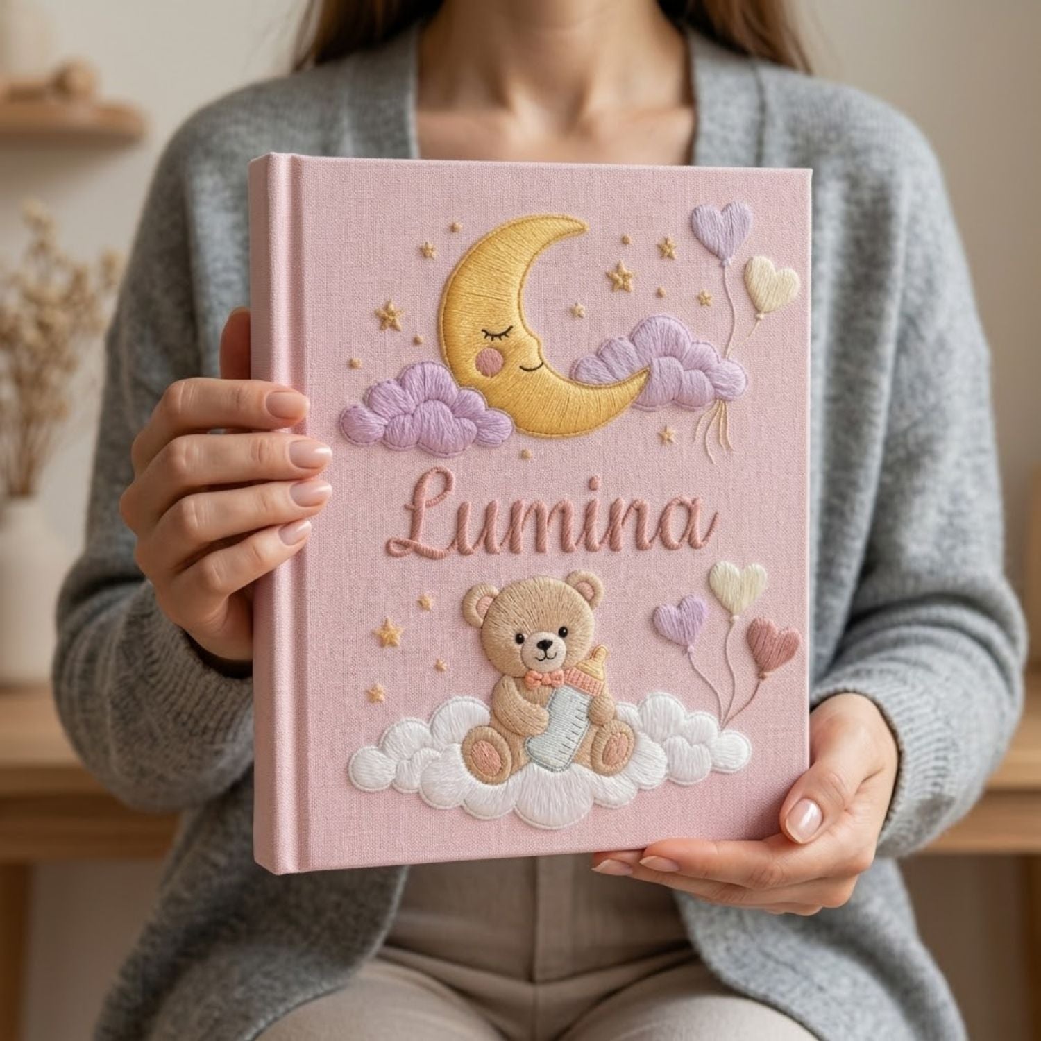 Personalized hand embroidered photo album featuring a teddy bear holding a baby bottle on fluffy clouds beneath a crescent moon with heart balloons and stars on a pink linen cover customized with a baby name