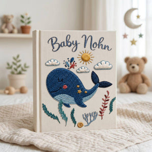 Personalized embroidered baby photo album with smiling whale, ocean plants, clouds, sunshine, and custom name embroidery